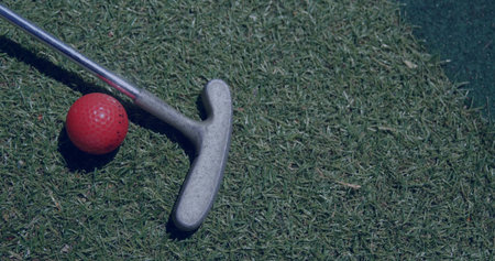 Resting silver golf putter head and red golf ball on trimmed putting green, with synthetic mat....