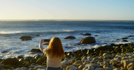 Capturing woman in sweater and jeans raising smartphone on beach at sunrise, framing ocean view. Seascape, tranquility, serenity, natural, outdoor, contemplative, minimalistの写真素材