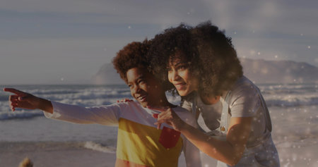 Leaning mother and son wearing denim overalls, color-blocked shirt pointing toward horizon on beach....