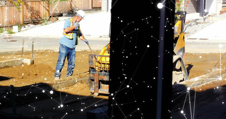 Using shovel, laborer in safety gear spreading soil beside loader at residential site, copy space. Construction, infrastructure, manual labor, development, urban, safety, outdoorの写真素材