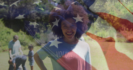 Smiling woman wearing VOLUNTEER T-shirt standing in park, team watering sapling with flag overlay. Community, environmentalism, teamwork, greenery, conservation, outdoors, empowermentの写真素材