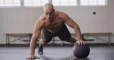 Shirtless man doing medicine-ball push-up on rubberized gym floor, with large windows. Fitness, strength, training, athleticism, workout, core, motivationの写真素材