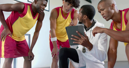Kneeling coach and players listening inside basketball gym, with tablet stopwatch jerseys and shoes. Sports, teamwork, training, athletics, fitness, motivation, performanceの写真素材