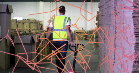 Walking worker wearing safety vest maneuvering pallet jack in warehouse aisle, with cardboard boxes....