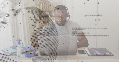 Working man wearing headset typing on laptop at home desk, with notebook, tablet and map overlay. Technology, remote work, digital connectivity, productivity, modern office, communication, innovationの写真素材