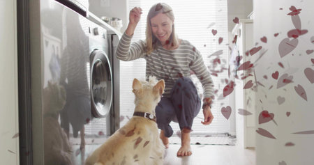 Holding treat, woman crouching in laundry area with front-load washer, dog collar, floating hearts. Pet, domestic, cozy, playful, caring, interaction, homeの写真素材