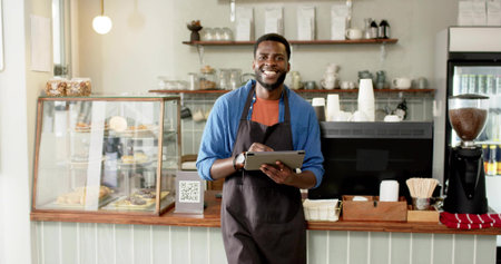 Smiling barista wearing blue shirt and apron holding tablet at cafe, with pastry display case. Coffee shop, hospitality, modern design, culinary, customer service, entrepreneurship, vibrant atmosphereの写真素材