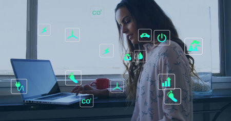 Woman in pink top typing at windowsill desk, with laptop mug floating energy icons, copy space. Environmentalist, feminine, modern, sustainable, innovative, cozy, creativeの写真素材