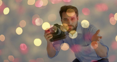 Crouching male photographer holding DSLR camera with zoom lens near face in studio bokeh backdrop. Portrait, creativity, vibrancy, art, professional, lighting, focusの写真素材