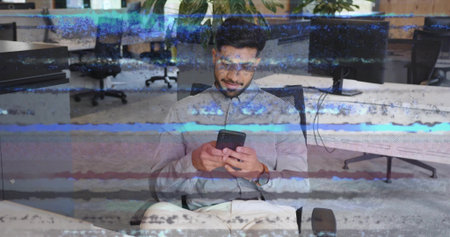 Indian office worker wearing glasses looking at smartphone sitting at desk in office, with monitors. Professional, productivity, contemporary, workspace, technology, focused, corporateの写真素材