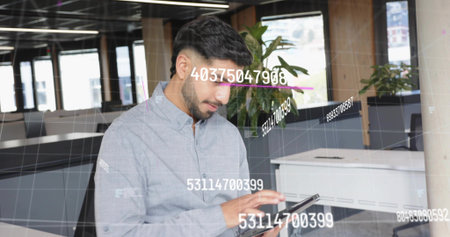 Interacting adult Indian man reviewing digital data overlays on tablet in open-plan office....