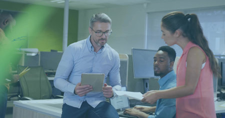 Male in suit holding tablet and leading discussion in open-plan office, with monitors and papers....