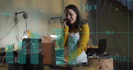 Speaking artisan woman holding smartphone and typing on laptop in workshop, with financial overlays. Craftsman, workspace, innovation, productivity, rustic, digital, entrepreneurshipの写真素材