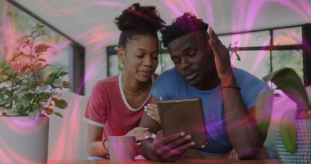 Browsing couple in red and blue shirts viewing tablet at home dining area, with mug, plant. Coziness, relaxation, domestic, modern, greenery, lifestyle, harmonyの写真素材