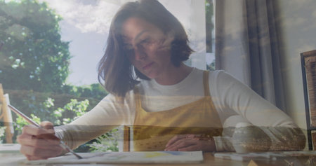 Adult female painter wearing yellow apron painting on paper at table by window, with paintbrush. Creative, tranquil, artistic, rustic, inspiring, leisurely, vibrantの写真素材
