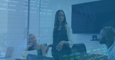 Presenting woman standing beside glass wall showing stock charts in meeting room, with laptops....