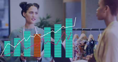 Women wearing knit sweater and blazer discussing sales in showroom, with clipboard chart. Fashion,...
