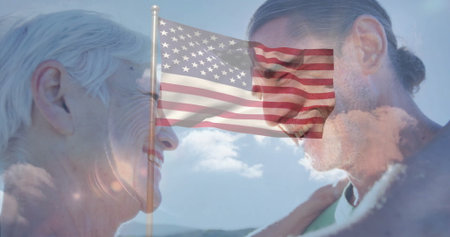 Smiling mother and son standing face to face on hilltop, with fluttering American flag on flagpole. Patriotism, family, generational, outdoor, serenity, unity, celebrationの写真素材