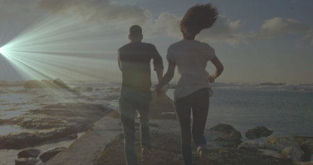 Running couple holding hands along stone jetty at sunset, with ocean tidal pools, scattered clouds....