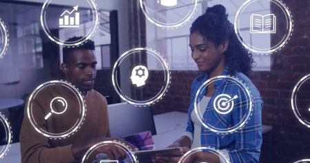 Gesturing male and female coworkers examining tablet near monitors at office, with floating icons. Collaboration, teamwork, innovation, digital, productivity, corporate, modernの写真素材