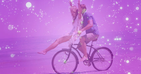 Pedaling barefoot couple balancing bicycle with woman raising arms on beach, with particle overlay. Adventure, freedom, leisure, serenity, nature, relaxation, outdoorの写真素材