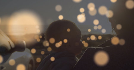 Mid adult woman holding infant boy at roadside at dusk, with parked sedan and bokeh flares. Family, maternal, tender, scenic, silhouette, affection, outdoorの写真素材