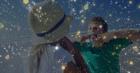 Adjusting straw hat, white-tee girl and green-tee boy pulling face at shoreline amid golden flecks. Children, beach, outdoor, playful, nature, leisure, happinessの写真素材