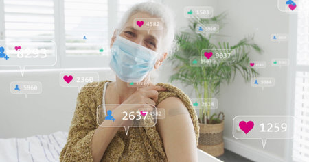 Displaying woman showing bandage wearing mask and sweater at home, with floating reaction icons. Health, wellness, elderly, immunity, social sharing, digital engagement, home environmentの写真素材