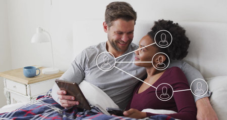 Using tablet and smartphone, couple wearing long-sleeve tops lounging on bed, with plaid blanket....