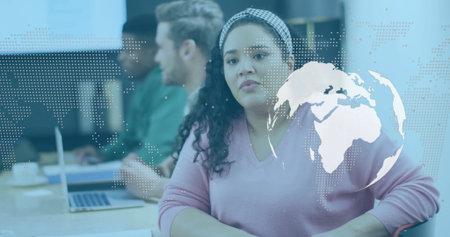Sitting Hispanic woman in pink sweater listening at conference table, laptops, globe overlay....
