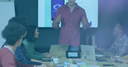 Presenting casually dressed man gesturing at head of conference table in meeting room, with display....