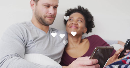 Couple using tablet and smartphone in living room on sofa, with pillow, blanket and floating hearts....