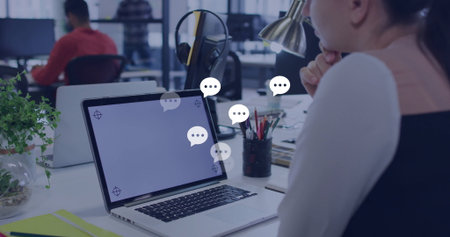 Focusing on laptop with chat bubbles, woman in vest in open office featuring terrarium....