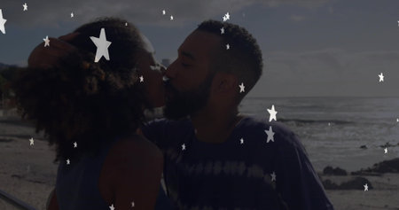 Kissing couple leaning close on sandy shoreline by railing with ocean waves, star-shaped overlays....