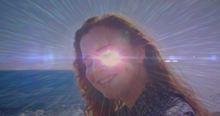 Smiling woman turning toward camera on coastline at sunset, with lens flare streaks over horizon. Vacation, serenity, outdoor, adventure, natural, vibrant, freedomの写真素材