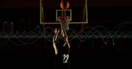 Shooting player wearing black number 22 jersey aiming ball toward hoop at gym, with light waves. Athlete, sport, modern, dynamic, competition, training, performanceの写真素材