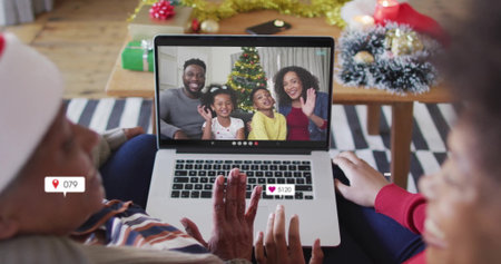 Showing women wearing Santa hat watching call on laptop in living room, with Christmas tree gifts. Family, connectivity, celebration, festivity, tradition, home decor, generationalの写真素材
