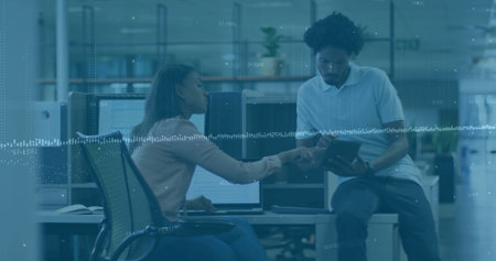 Pointing woman guiding coworker through tablet data in modern open-plan office, with dual monitors....