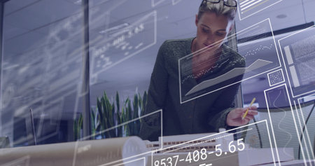 Examining architect in blouse leaning over scale model at modern studio, with monitors showing code. Creativity, innovation, collaboration, productivity, technical, professional, minimalisticの写真素材