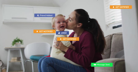 Holding mother leaning forward kissing baby cheek on couch at home, with notification overlays. Family, connection, domestic, contemporary, communication, affectionate, leisureの写真素材