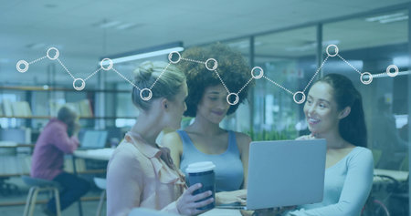 Collaborating women pointing at laptop holding coffee cups in modern workspace at high table....