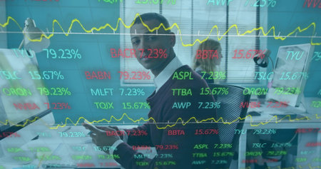 South Asian man wearing business casual holding device watching stock tickers at desk with keyboard....