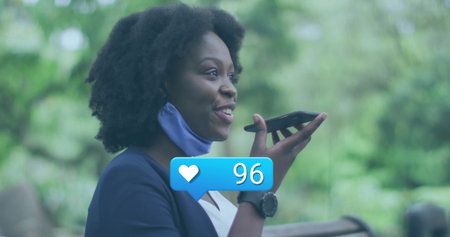 Woman wearing blazer, smartwatch, mask on chin, using smartphone on bench in park, with like icon. Outdoor, communication, technology, leisure, wellness, casual, natureの写真素材