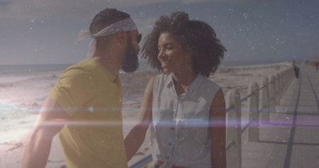 Walking couple wearing bandana, sunglasses, denim top on boardwalk planks by sea with metal railing....