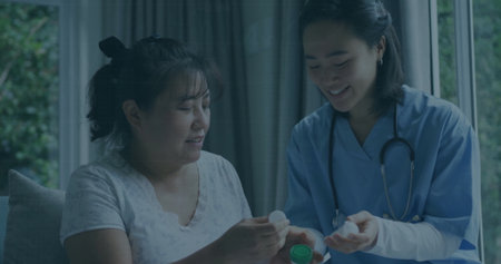 Holding pill bottle, nurse in blue scrubs, stethoscope guiding patient examining cap in living room. Healthcare, wellness, medical, caregiving, home, compassion, tranquilityの写真素材