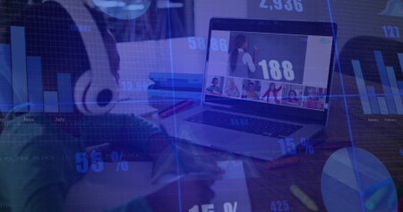 Writing user wearing headphones noting in notebook, watching laptop at desk with data overlays. Technology, remote work, productivity, digital analysis, concentration, communication, home officeの写真素材