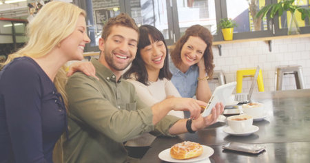 Chatting friends lifting coffee cups and viewing tablet at modern cafe, with pastry and plants. Socializing, collaboration, urban, contemporary, cozy, leisure, engagementの写真素材