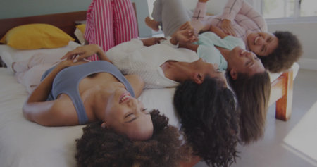 Laughing four friends wearing pajamas lying on bed in bedroom, with white linens and yellow pillows. Cozy, friendship, relaxation, leisure, comfort, wellness, familialの写真素材