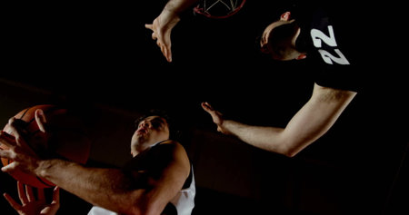 Jumping two male basketball players battling under hoop in sports hall, with basketball and jerseys....