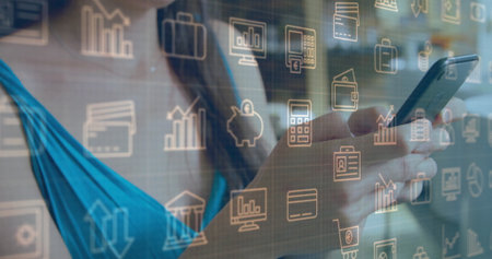 Holding smartphone, man wearing turquoise top viewing financial tech icons at office desk. Technology, finance, digital, professional, modern, innovation, dataの写真素材
